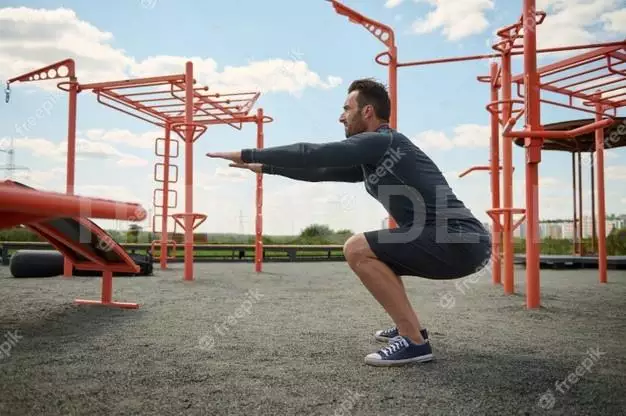 Active Athlete In Athletic Uniform Performing Squats During Training On An Outdoor Sports Field. Young Man Doing Sports On The Summer Sportsground. Healthy And Active Lifestyle Concept 