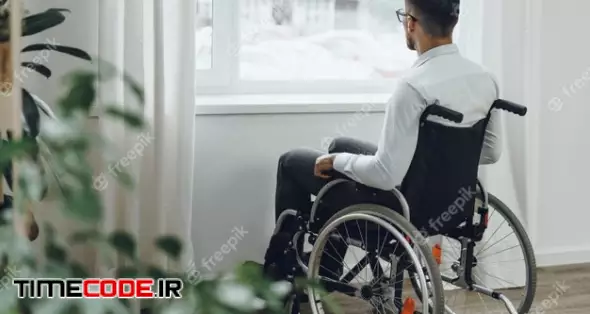 Young Disabled Man Sitting In A Wheelchair Near The Window At Home 