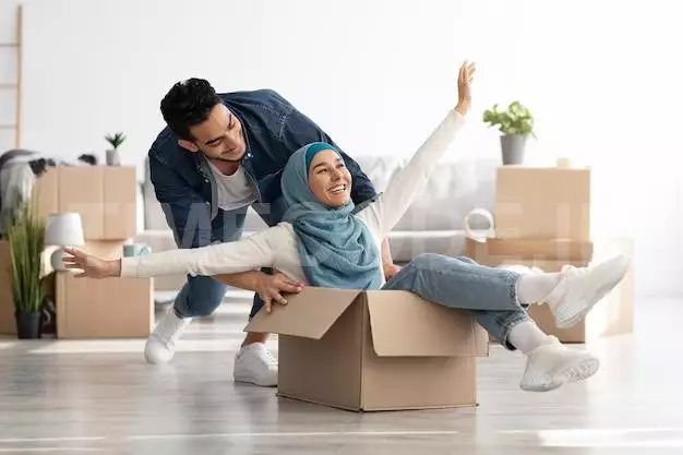 Joyful Middle-eastern Young Family Having Fun While Unpacking Stuff In Their New House. Happy Arab Guy And Excited Lady In Hijab Sitting In Paper Box, Enjoying Moving. Apartment, House, Real Estate