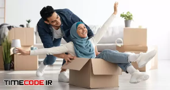 Joyful Middle-eastern Young Family Having Fun While Unpacking Stuff In Their New House. Happy Arab Guy And Excited Lady In Hijab Sitting In Paper Box, Enjoying Moving. Apartment, House, Real Estate