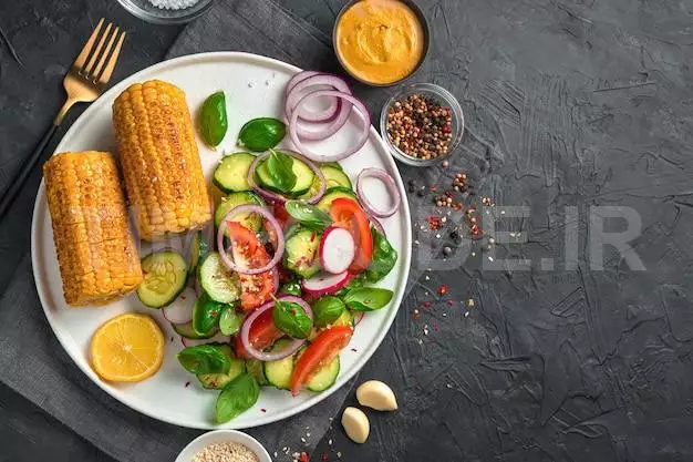 Vegetable Salad With Tomatoes, Cucumbers And Radishes And Heads Of Corn In A White Flat Plate On A Black Wall With Space To Copy. The Concept Of Vegetarian Food.