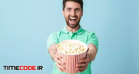 Portrait Of A Handsome Bearded Man Wearing Casual Clothing Standing Isolated Over Blue Wall, Eating Popcorn While Watching Movie